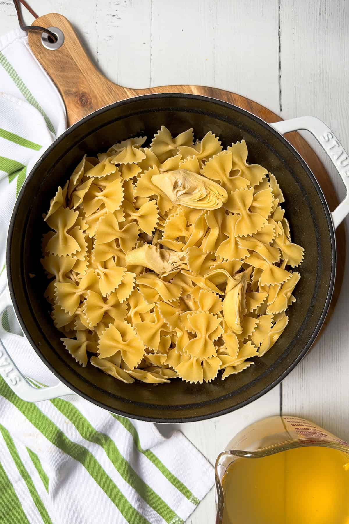 A black pot filled with uncooked farfalle, artichoke hearts, and ingredients for a lemon asparagus pasta sits on a wooden board next to a striped kitchen towel and a pitcher of broth.