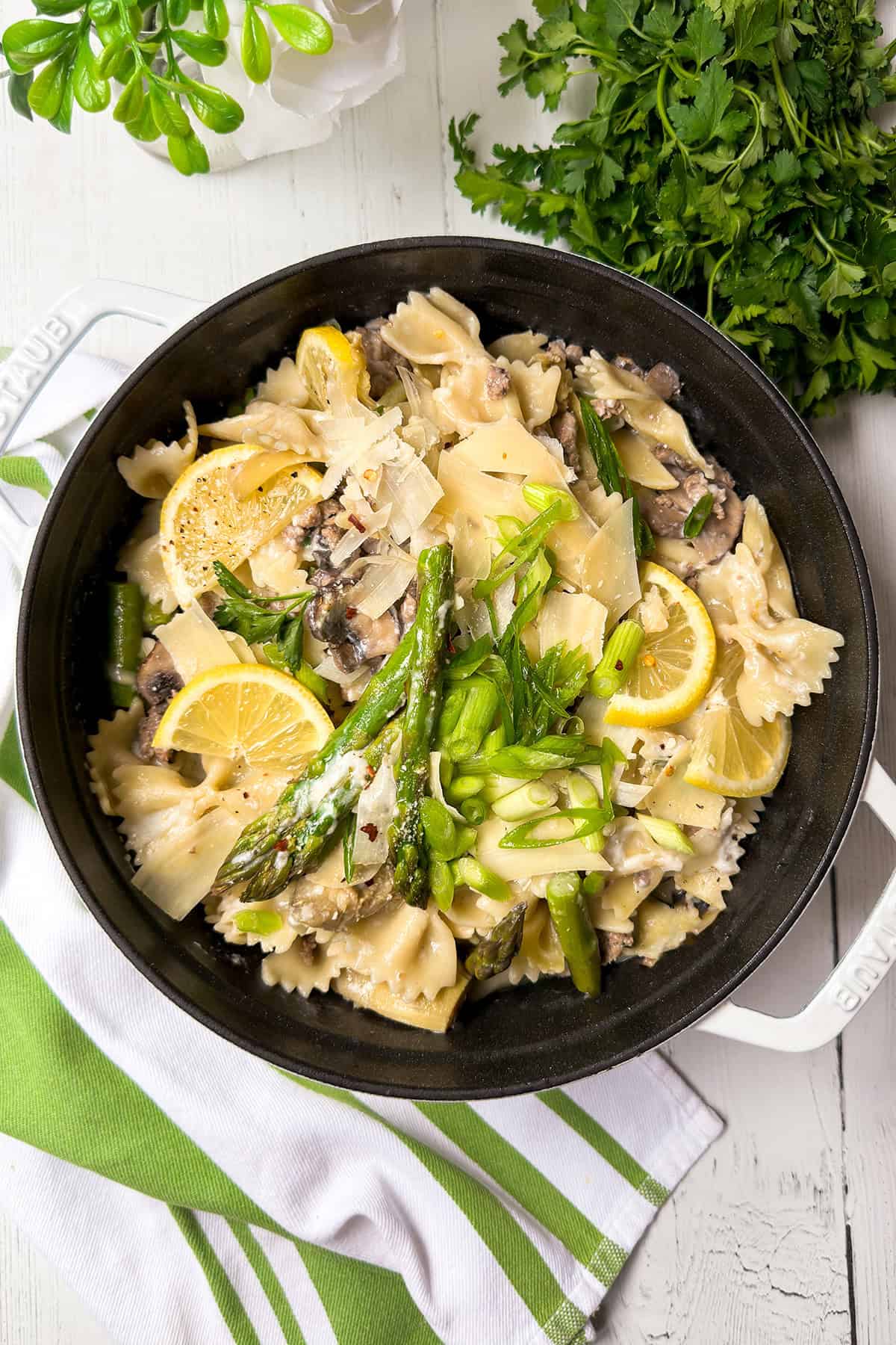 A black pot filled with creamy asparagus pasta, farfalle, lemon slices, mushrooms, and parmesan sits on a white table next to fresh parsley and a green-striped towel-perfect for your next asparagus pasta recipe.