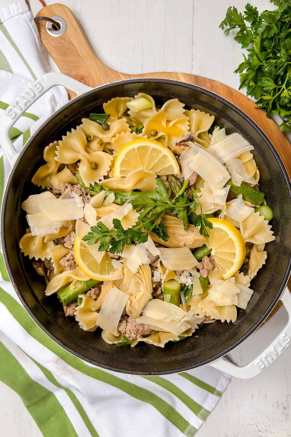 A black pot filled with lemon asparagus pasta, ground meat, parsley, and shaved cheese sits on a wooden board next to a bunch of fresh parsley.
