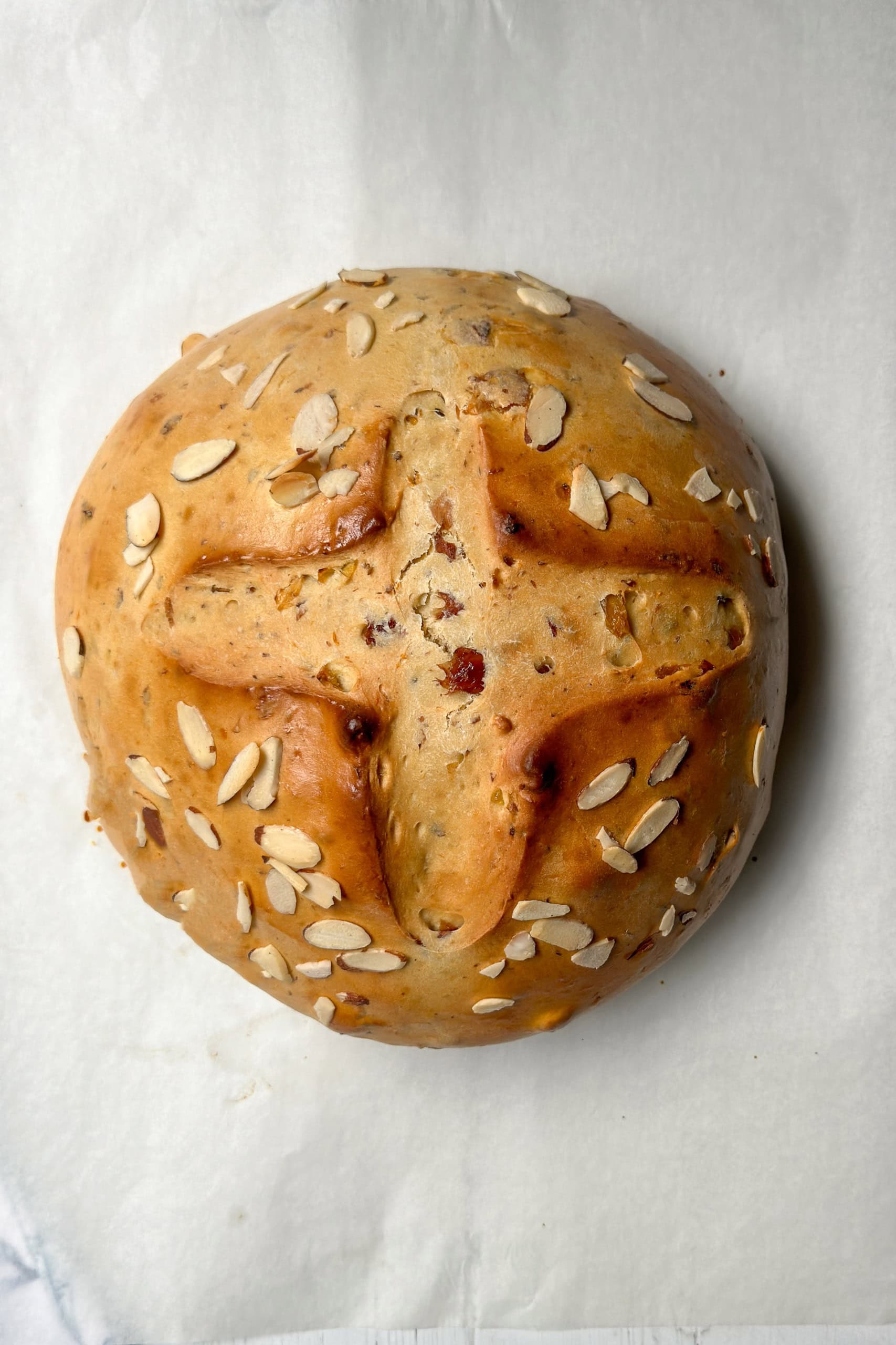 A round loaf of German Easter Bread topped with sliced almonds, featuring a cross-shaped slash on top, rests on a sheet of parchment paper.