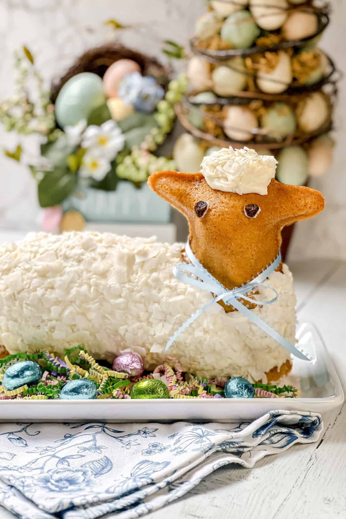 A delightful Easter lamb cake decorated with white frosting, a blue ribbon, and chocolate eyes, surrounded by Easter eggs and festive decorations.