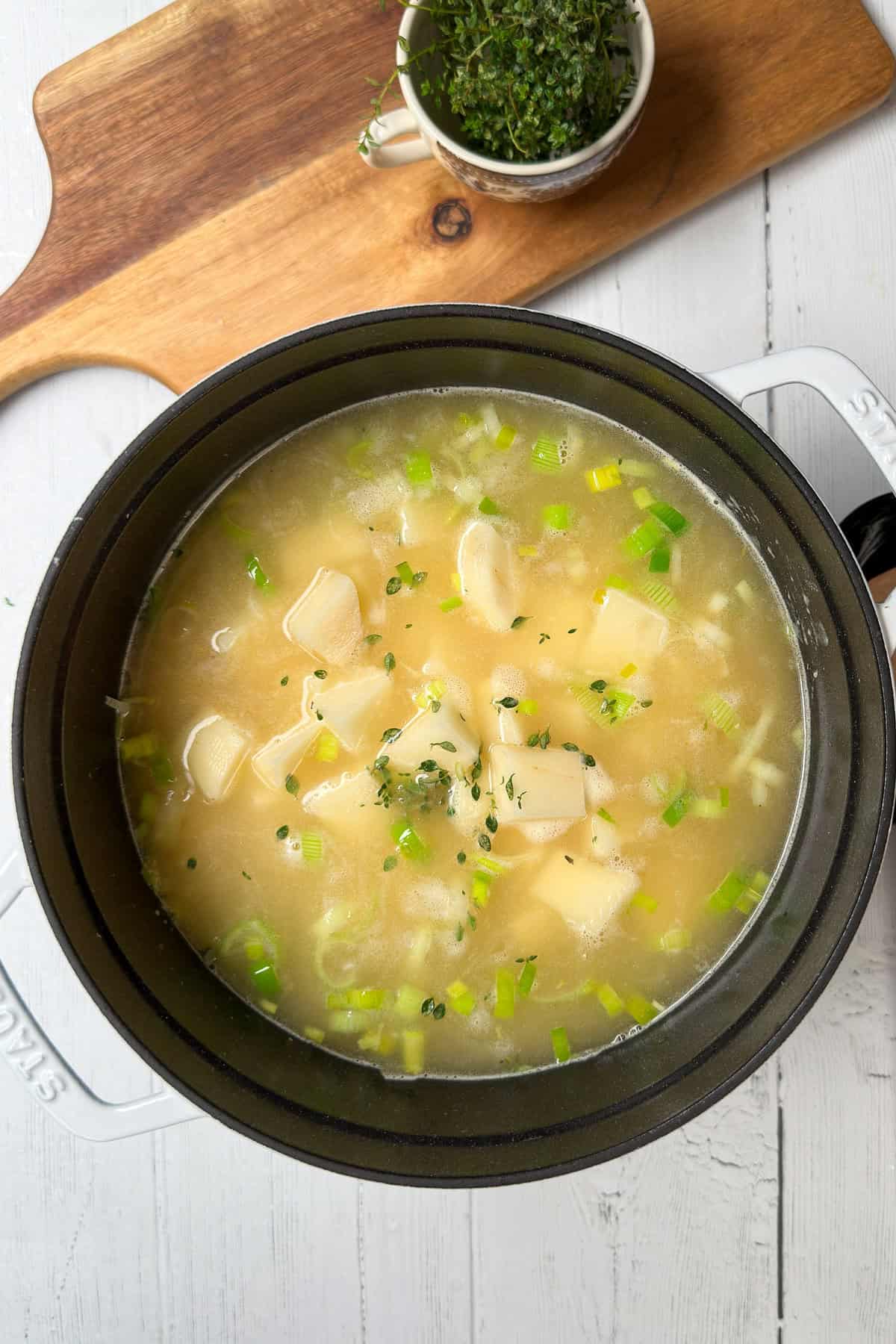 Top view of potatoes added to potato leek soup in a white Dutch oven with fresh thyme.