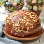 A round loaf of German Easter Bread topped with sliced almonds sits on a wooden plate, with decorative eggs and greenery in the background.