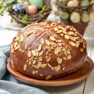 A round loaf of German Easter Bread topped with sliced almonds sits on a wooden plate, with decorative eggs and greenery in the background.