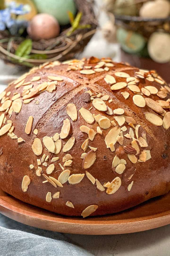 A round loaf of German Easter Bread topped with sliced almonds sits on a wooden plate, with decorative eggs and greenery in the background.