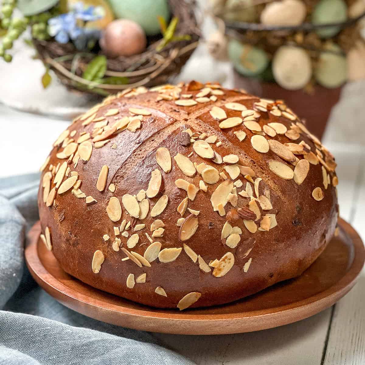 A round loaf of German Easter Bread topped with sliced almonds sits on a wooden plate, with decorative eggs and greenery in the background.