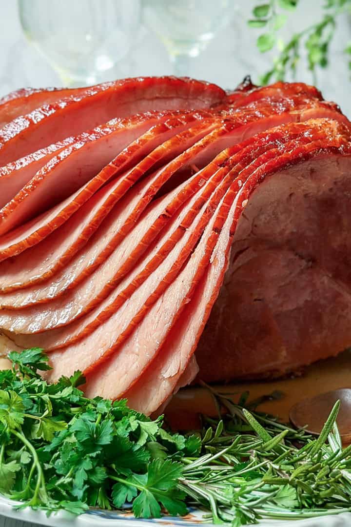 Sliced honey baked ham served on a platter with fresh parsley and rosemary garnishes.