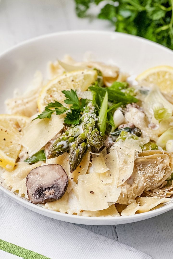A bowl of lemon asparagus pasta with mushrooms, lemon slices, grated cheese, and parsley, set on a white table with a green herb garnish in the background.