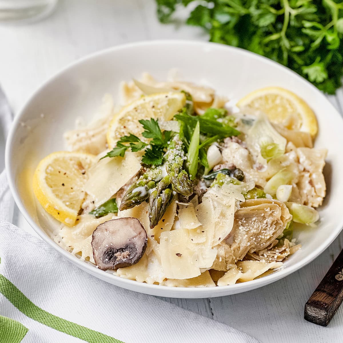 A bowl of lemon asparagus pasta with mushrooms, lemon slices, grated cheese, and parsley, set on a white table with a green herb garnish in the background.