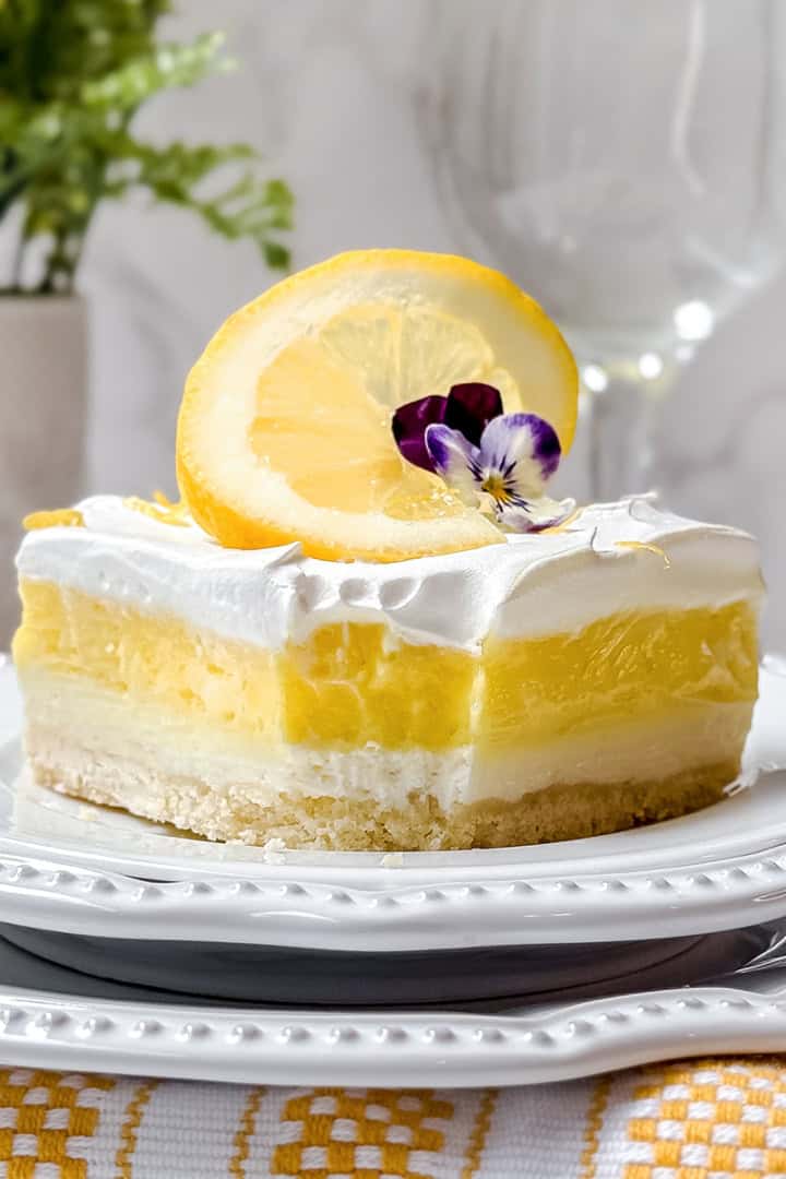 A slice of layered lemon dessert with whipped topping, garnished with a lemon slice and edible flowers, served on a white plate-perfect for Easy Easter Desserts.