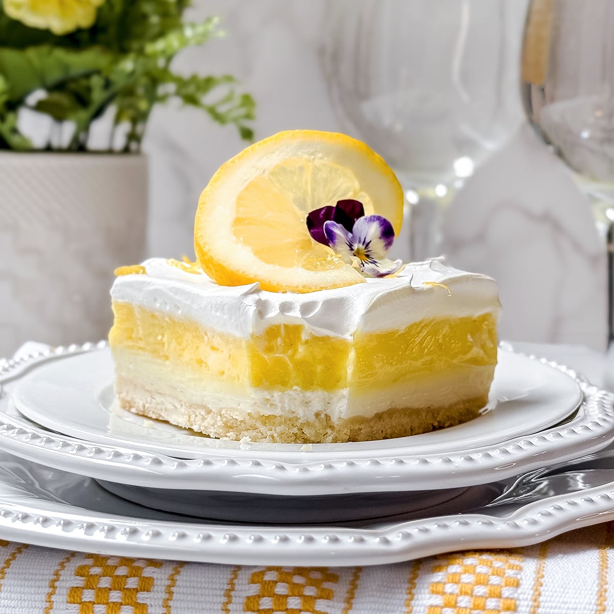 A slice of layered lemon dessert with whipped topping, garnished with a lemon slice and edible flowers, served on a white plate—perfect for Easy Easter Desserts.