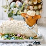 Easter lamb cake decorated with white frosting and a blue ribbon, surrounded by colorful Easter eggs and artificial grass on a white platter.