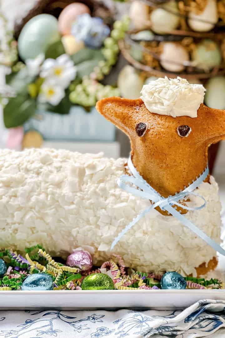 Easter lamb cake decorated with white frosting and a blue ribbon, surrounded by colorful Easter eggs and artificial grass on a white platter.