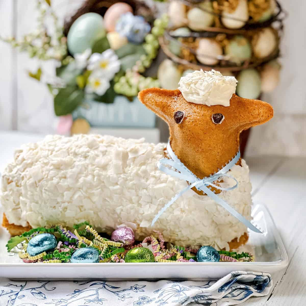 Easter lamb cake decorated with white frosting and a blue ribbon, surrounded by colorful Easter eggs and artificial grass on a white platter.