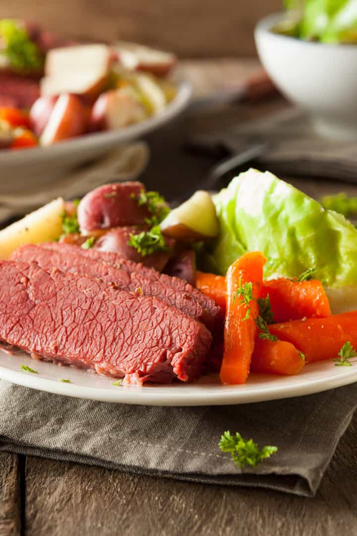 A plate with slices of corned beef, boiled carrots, potatoes, and a wedge of cabbage, garnished with parsley, sits on a rustic wooden table.