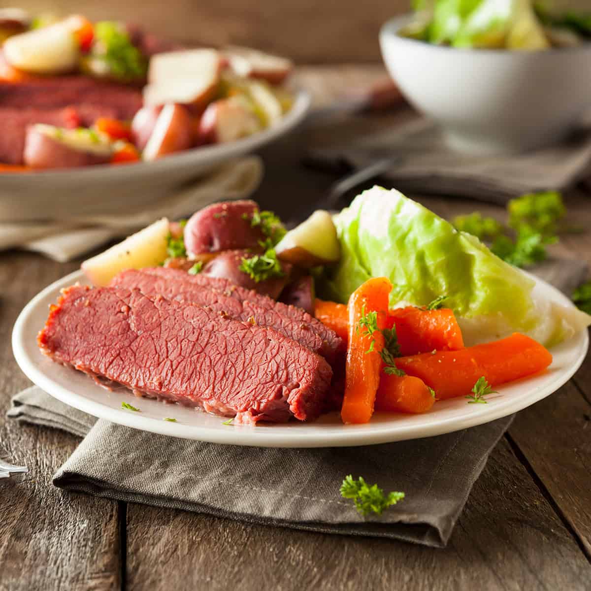 A plate with slices of corned beef, boiled carrots, potatoes, and a wedge of cabbage, garnished with parsley, sits on a rustic wooden table.