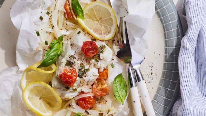 A baked fish fillet with cherry tomatoes, onion slices, lemon slices, and basil leaves served on parchment paper—an ideal choice for March One Pot Dinners—presented on a plate with a fork and knife.