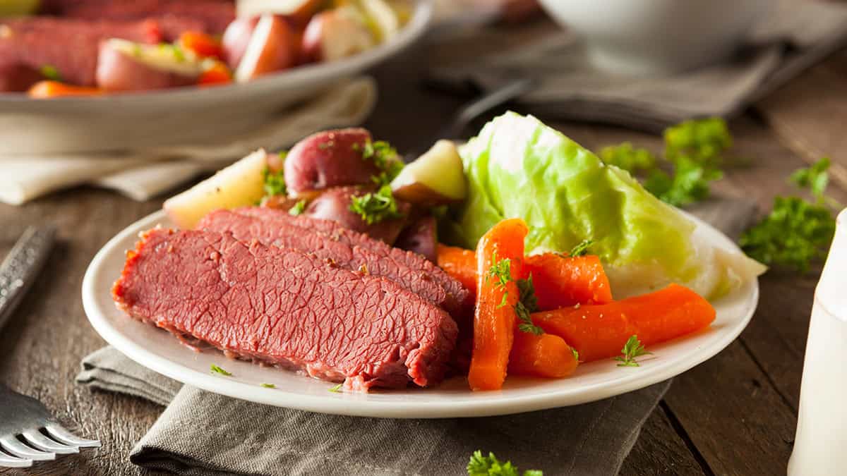 A plate of savory corned beef with boiled carrots, potatoes, and cabbage, garnished with parsley, sits on a wooden table with a napkin underneath.