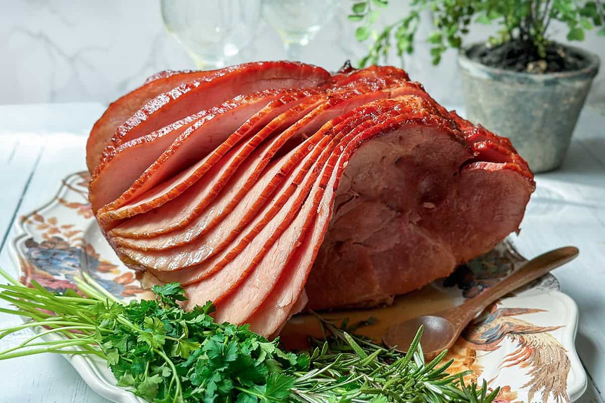A honey baked ham, spiral-sliced and glistening, sits on a decorative platter garnished with fresh parsley and rosemary, with a wooden spoon and potted plant in the background.