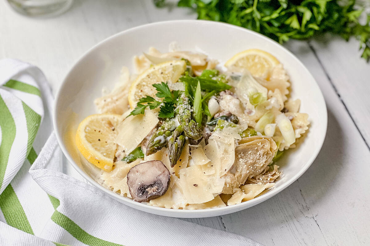 A white bowl of lemon asparagus pasta with mushrooms, artichokes, lemon slices, and fresh parsley sits on a white wooden surface.