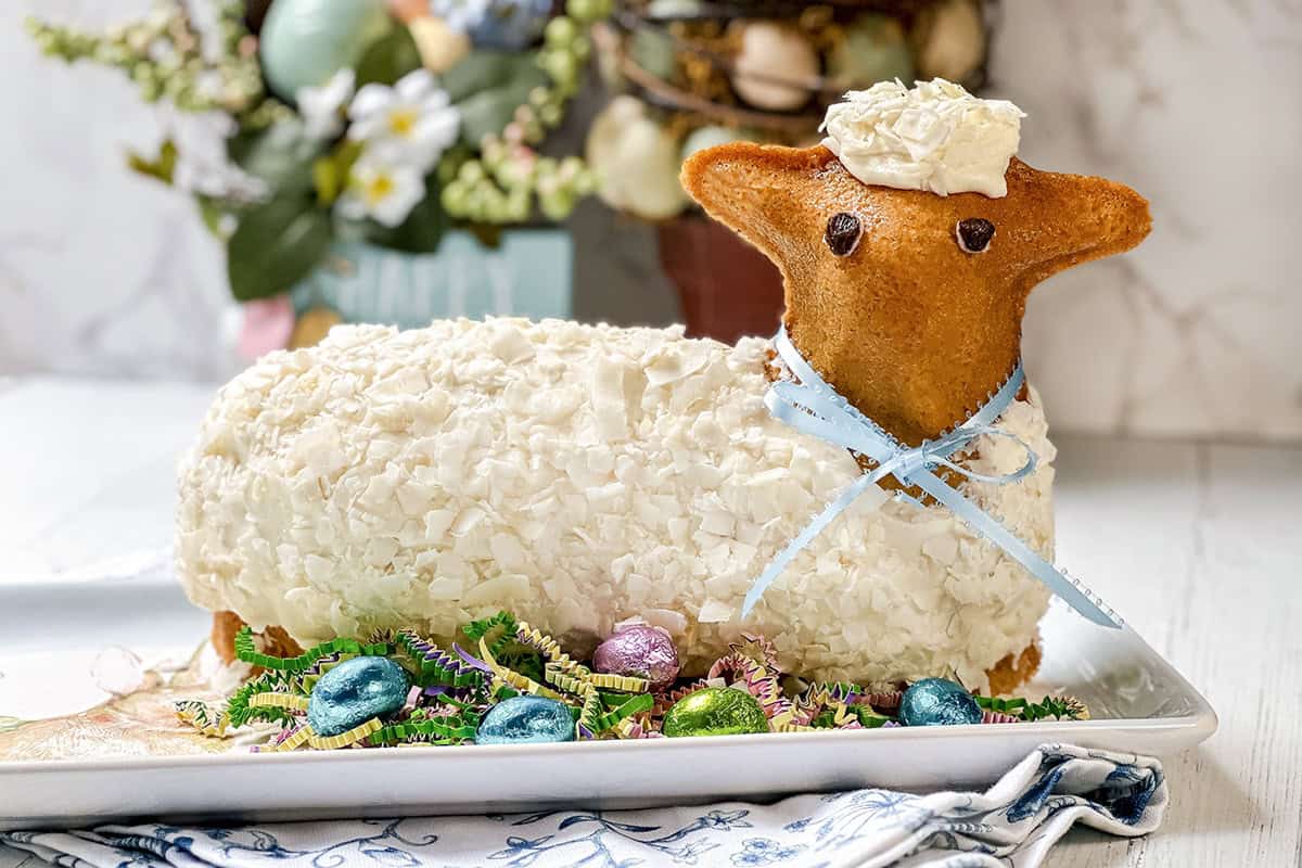 A festive Easter lamb cake covered in white frosting and coconut flakes, with chocolate eyes, a blue ribbon collar, and surrounded by colorful Easter eggs and decorations.
