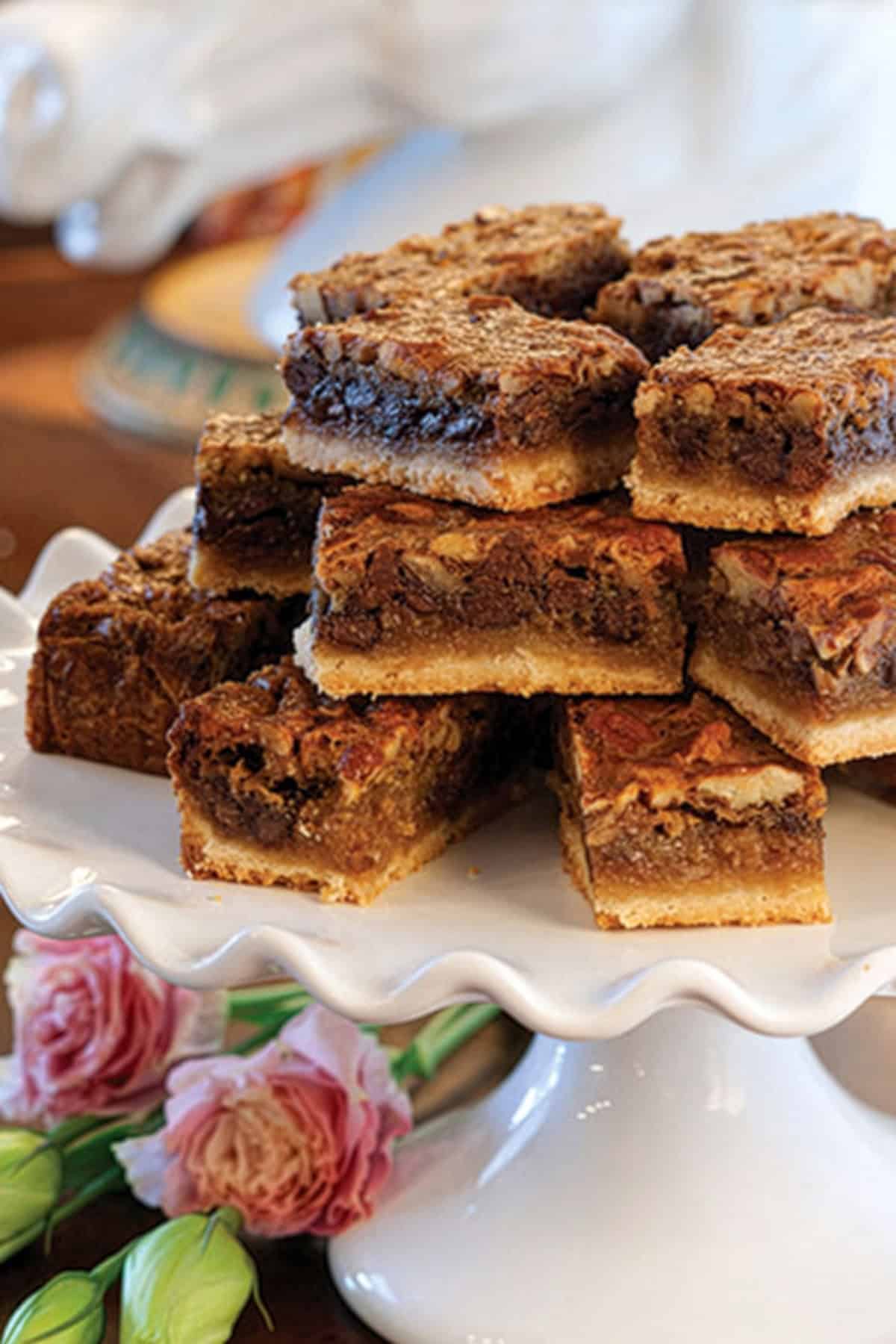 A stack of Kentucky Derby Bars with a golden crust displayed on a white scalloped cake stand, with pink flowers in the foreground.