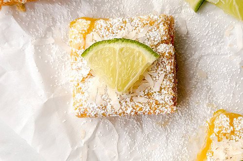 Lime bars topped with powdered sugar, shredded coconut, and lime wedges are arranged on white parchment paper alongside slices of fresh lime.