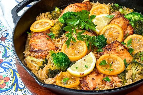 Side view of Lemon Chicken in a black cast iron skillet with broccoli, lemon slices, and orzo.