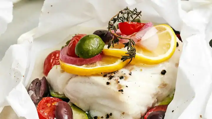 A piece of white fish topped with lemon, herbs, and vegetables sits on parchment paper, surrounded by olives, tomatoes, and sliced onions—a perfect choice for April Mediterranean Dinners.