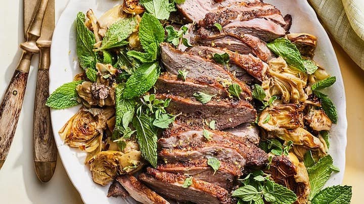 Distant view of sliced lamb roast with artichokes and mint leaves in a white oval serving dish.