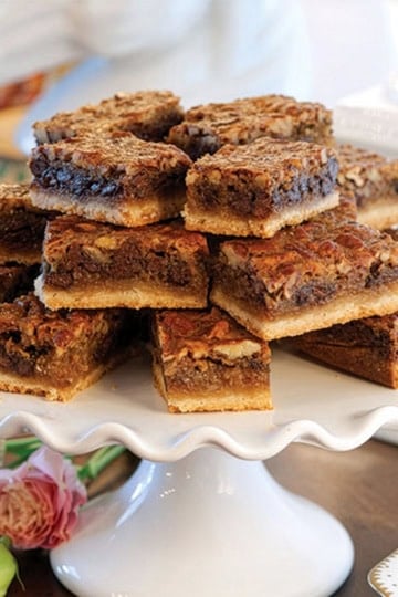 A stack of Kentucky Derby Bars with a cookie crust is arranged on a white pedestal cake stand. Pink flowers are placed nearby.