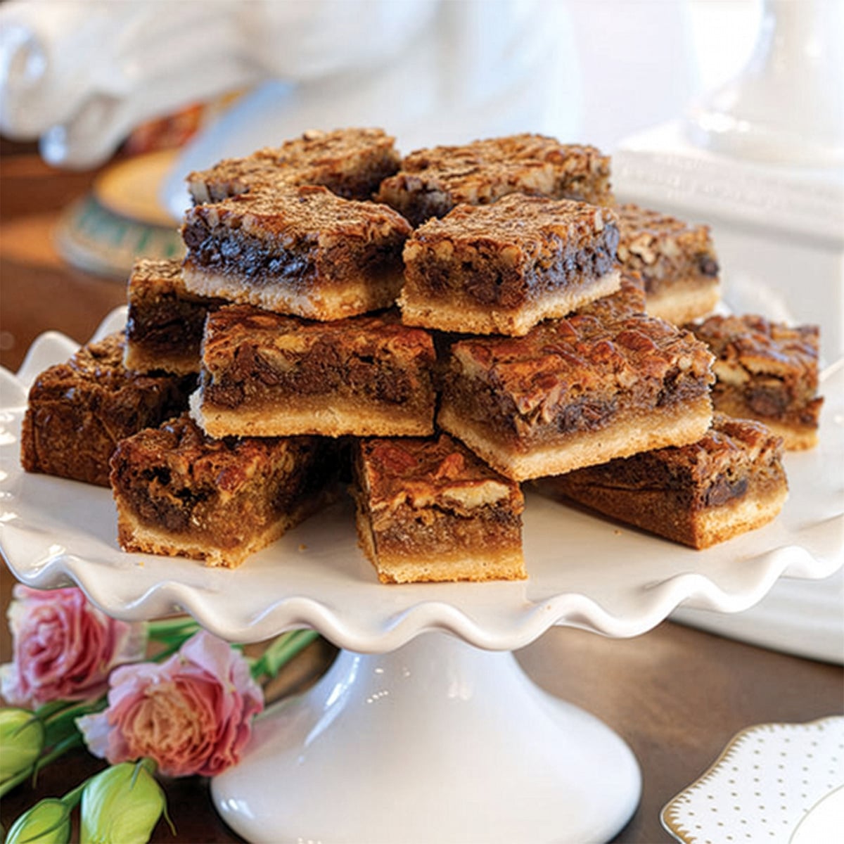 A stack of Kentucky Derby Bars with a cookie crust is arranged on a white pedestal cake stand. Pink flowers are placed nearby.