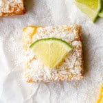 A square lime bar topped with shredded coconut, powdered sugar, and a lime wedge sits on white parchment paper.
