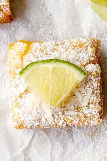 A square lime bar topped with shredded coconut, powdered sugar, and a lime wedge sits on white parchment paper.