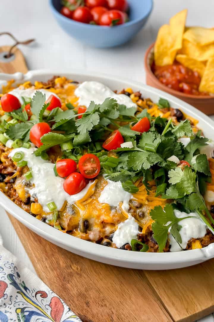Taco casserole in a large oval baking dish with tortillas, cherry tomatoes, and salsa on the side.