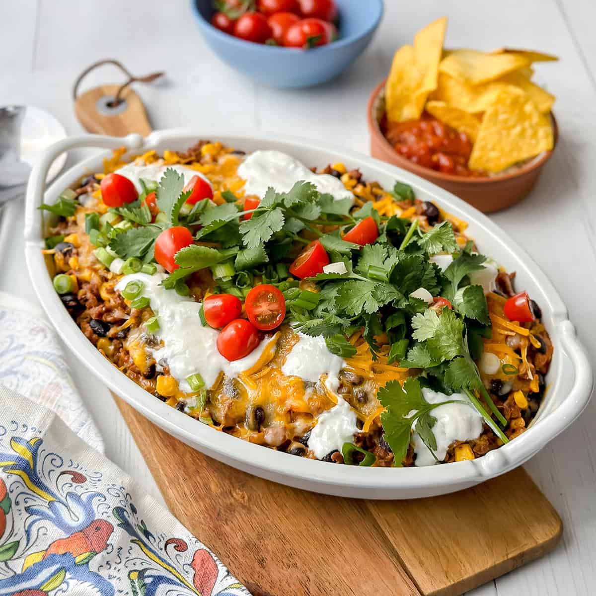 Taco casserole in a large oval baking dish with tortillas, cherry tomatoes, and salsa on the side.