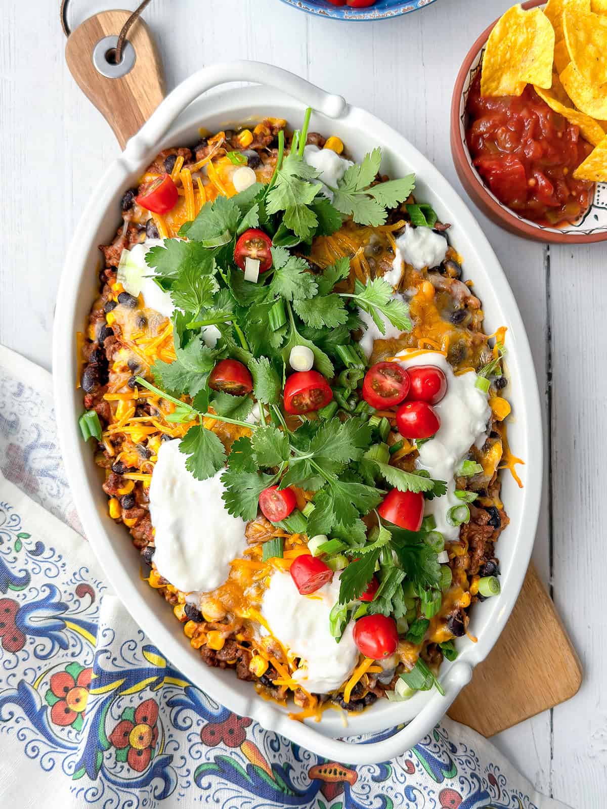 A savory taco casserole topped with melted cheese, sour cream, cilantro, chopped tomatoes, and green onions, served beside a bowl of salsa and tortilla chips on a patterned tablecloth.
