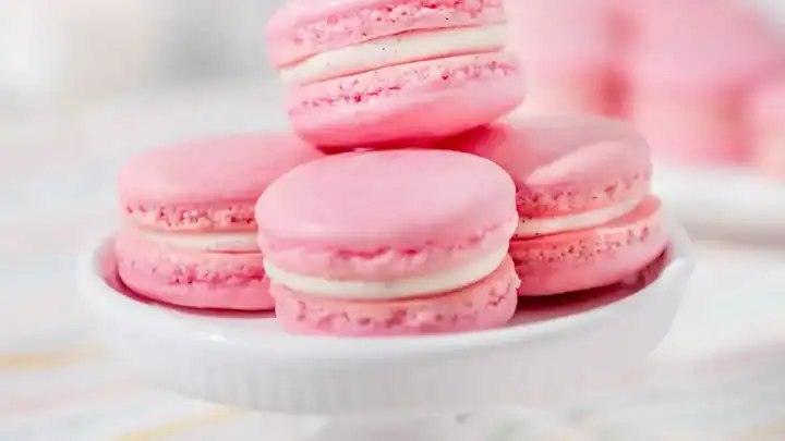 A small white cake stand displays five pink macarons with cream filling, set on a striped cloth—perfect for a charming bridal tea party.