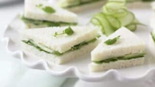 Triangular cucumber tea sandwiches garnished with microgreens are arranged on a white pedestal plate, with ribboned cucumber slices in the center—perfect for an elegant bridal tea party.