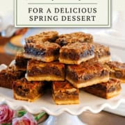 A stack of Kentucky Derby Bars is displayed on a white cake stand with pink flowers nearby, accompanied by a sign reading "Kentucky Derby Bars"—the perfect treat for a delicious spring dessert.