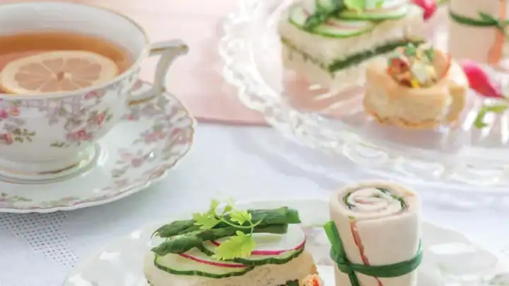 A plate of assorted tea sandwiches and pastries with a cup of tea and a lemon slice, all on floral china atop a pastel table setting—perfect for a charming bridal tea party.