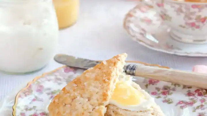 A scone with cream and lemon curd on a floral plate, accompanied by a butter knife, a jar of cream, and a cup of tea in a floral teacup—perfect for an elegant bridal tea party setting.