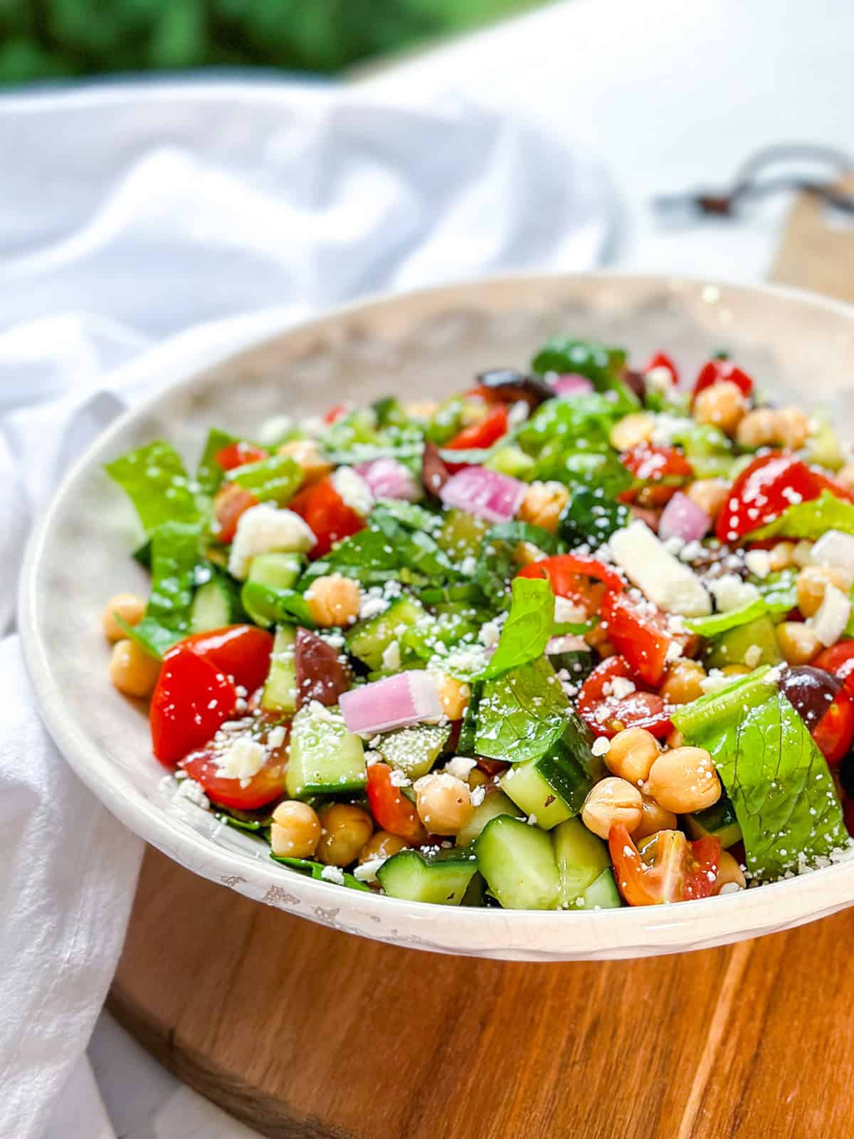 A bowl of Mediterranean Chopped Salad with chickpeas, chopped cucumbers, cherry tomatoes, red onions, spinach, and crumbled cheese sits on a wooden board.
