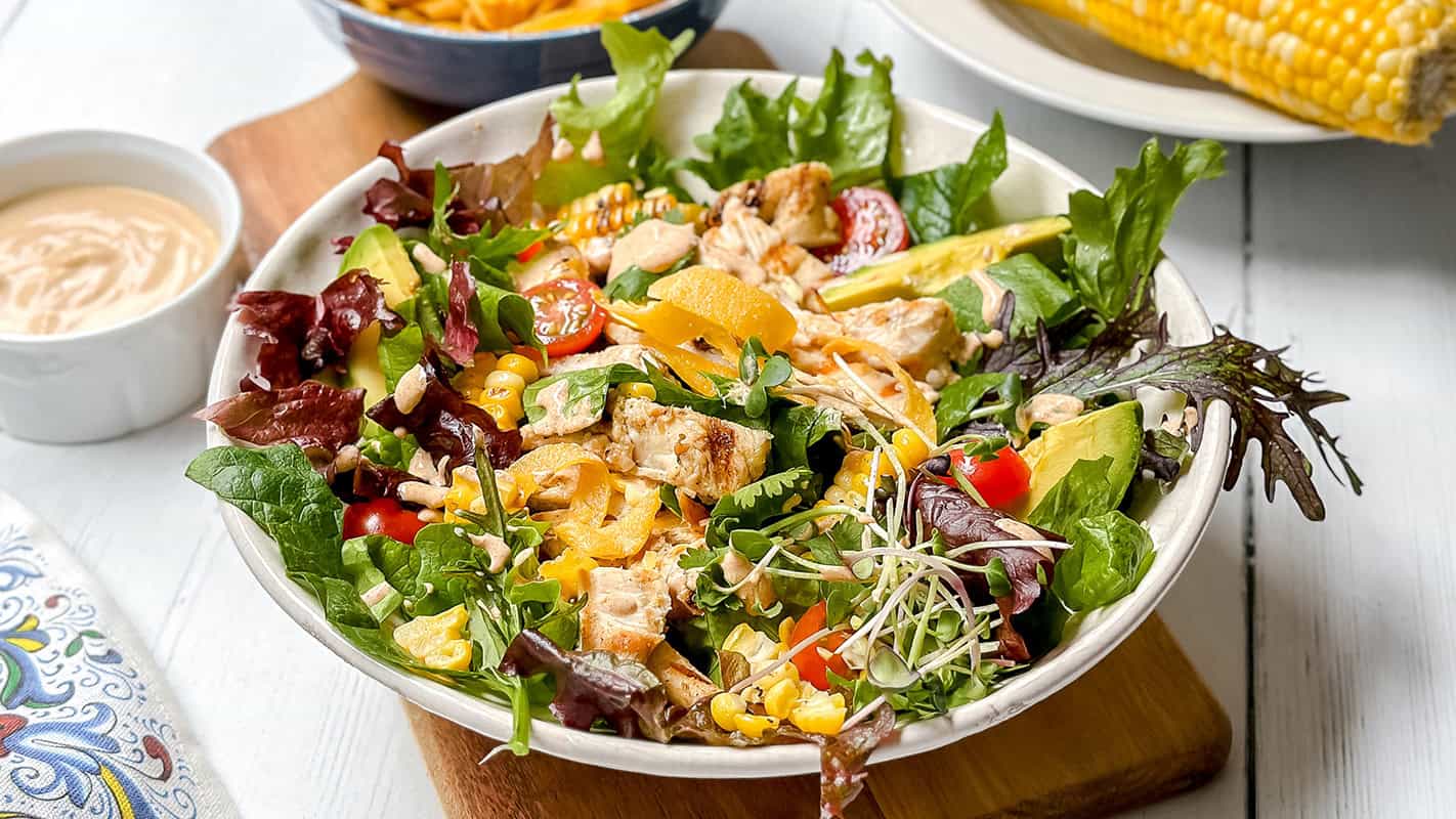 A vibrant southwest chicken salad featuring mixed greens, grilled chicken, sliced avocado, cherry tomatoes, bell peppers, corn, sunflower seeds, and sprouts. A side of dressing and corn on the cob accompany this fresh bowl.