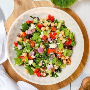 A plate of Mediterranean Chopped Salad with lettuce, cherry tomatoes, cucumber, red onion, chickpeas, black olives, and feta cheese on a wooden board.