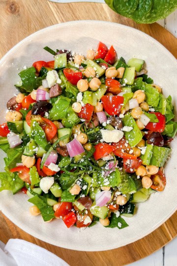 A plate of Mediterranean Chopped Salad with lettuce, cherry tomatoes, cucumber, red onion, chickpeas, black olives, and feta cheese on a wooden board.