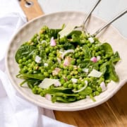 Bowl of pea salad with green peas, spinach, chopped onions, shaved cheese, and dressing, served with metal tongs on a wooden surface.