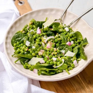 Bowl of pea salad with green peas, spinach, chopped onions, shaved cheese, and dressing, served with metal tongs on a wooden surface.