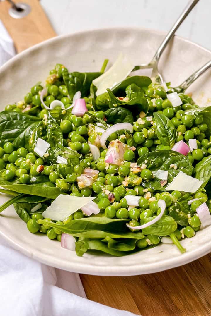 Bowl of pea salad with green peas, spinach, chopped onions, shaved cheese, and dressing, served with metal tongs on a wooden surface.