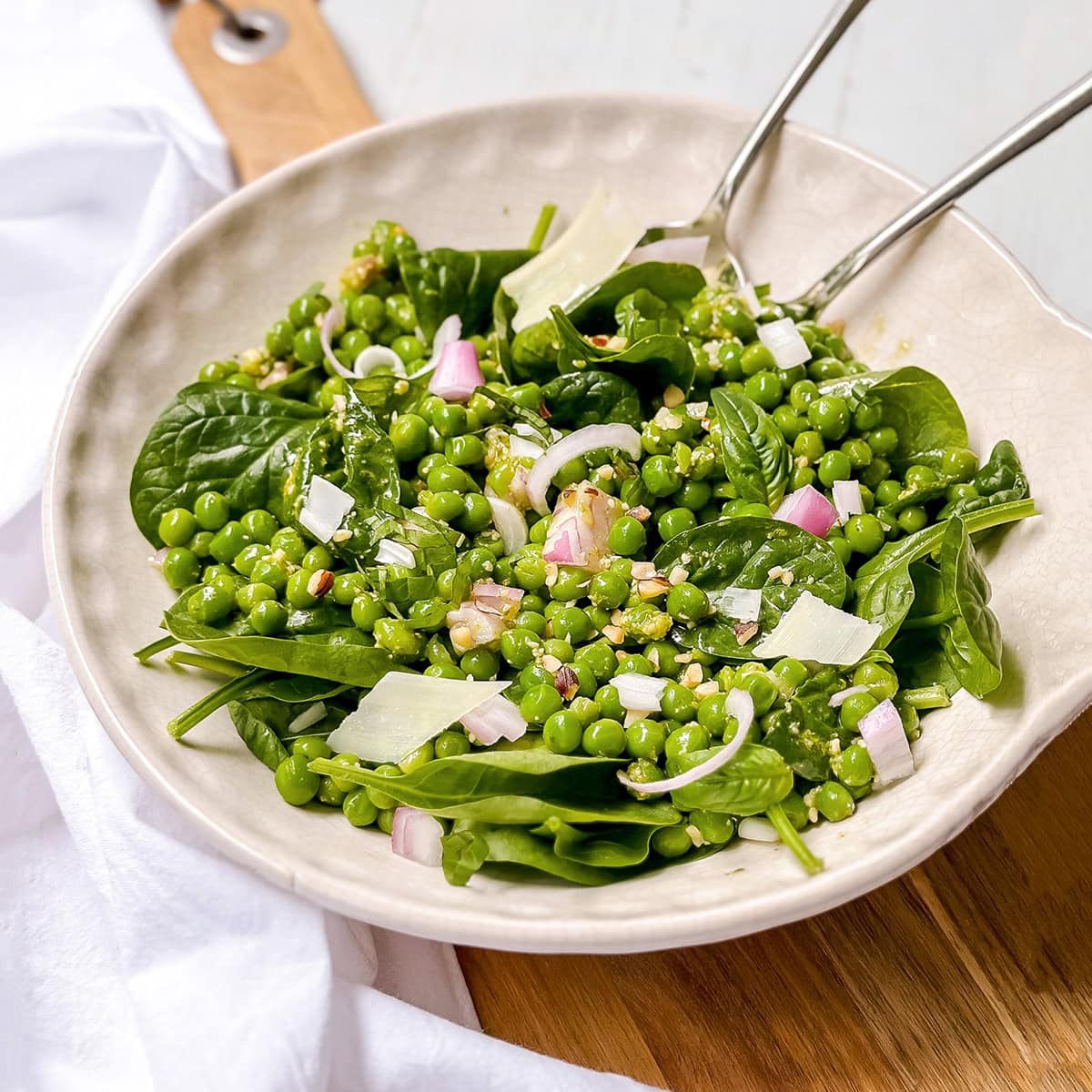 Bowl of pea salad with green peas, spinach, chopped onions, shaved cheese, and dressing, served with metal tongs on a wooden surface.